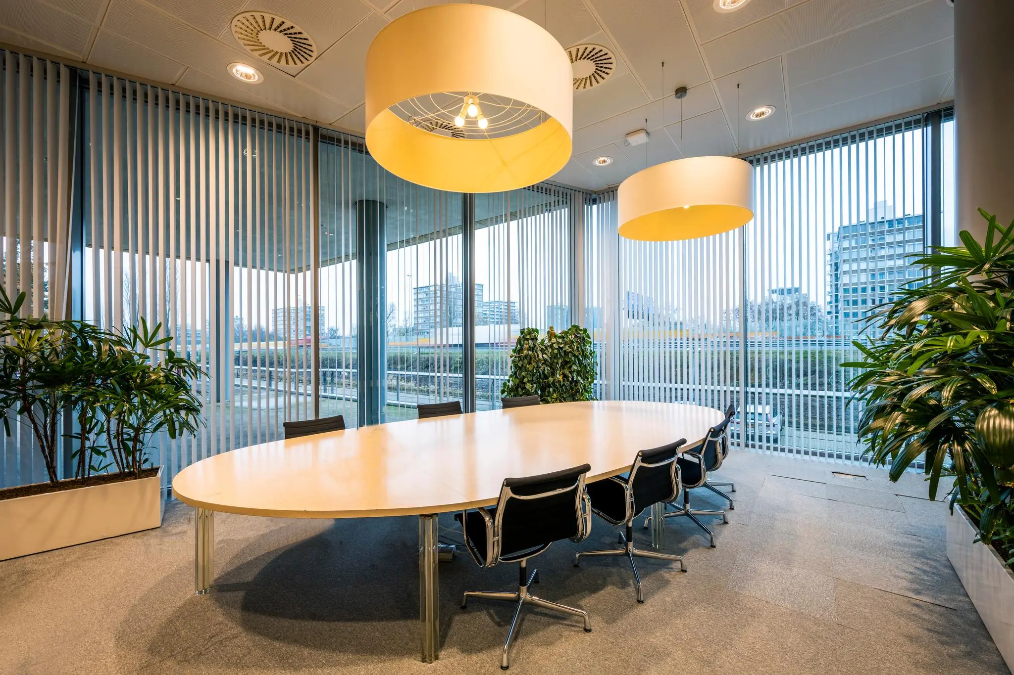 Moderne vergaderruimte aan de Burgemeester Rijnderslaan met grote ovale tafel, designstoelen, hanglampen en veel daglicht door hoge ramen met verticale lamellen.